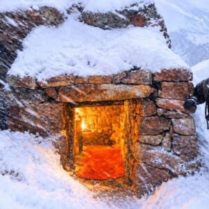 Stone survival shelter built under a giant rock overhang during a heavy mountain snowstorm