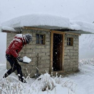 Stone survival shelter with a snow-covered roof nestled among snow-covered pine trees in a winter forest
